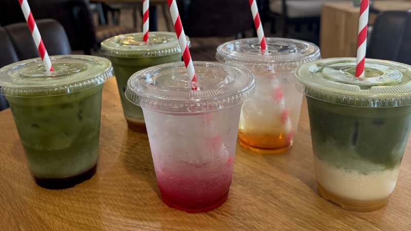 Five iced drinks in plastic cups with red-and-white striped straws lined up on a wooden table, including green matcha lattes and colorful iced beverages.